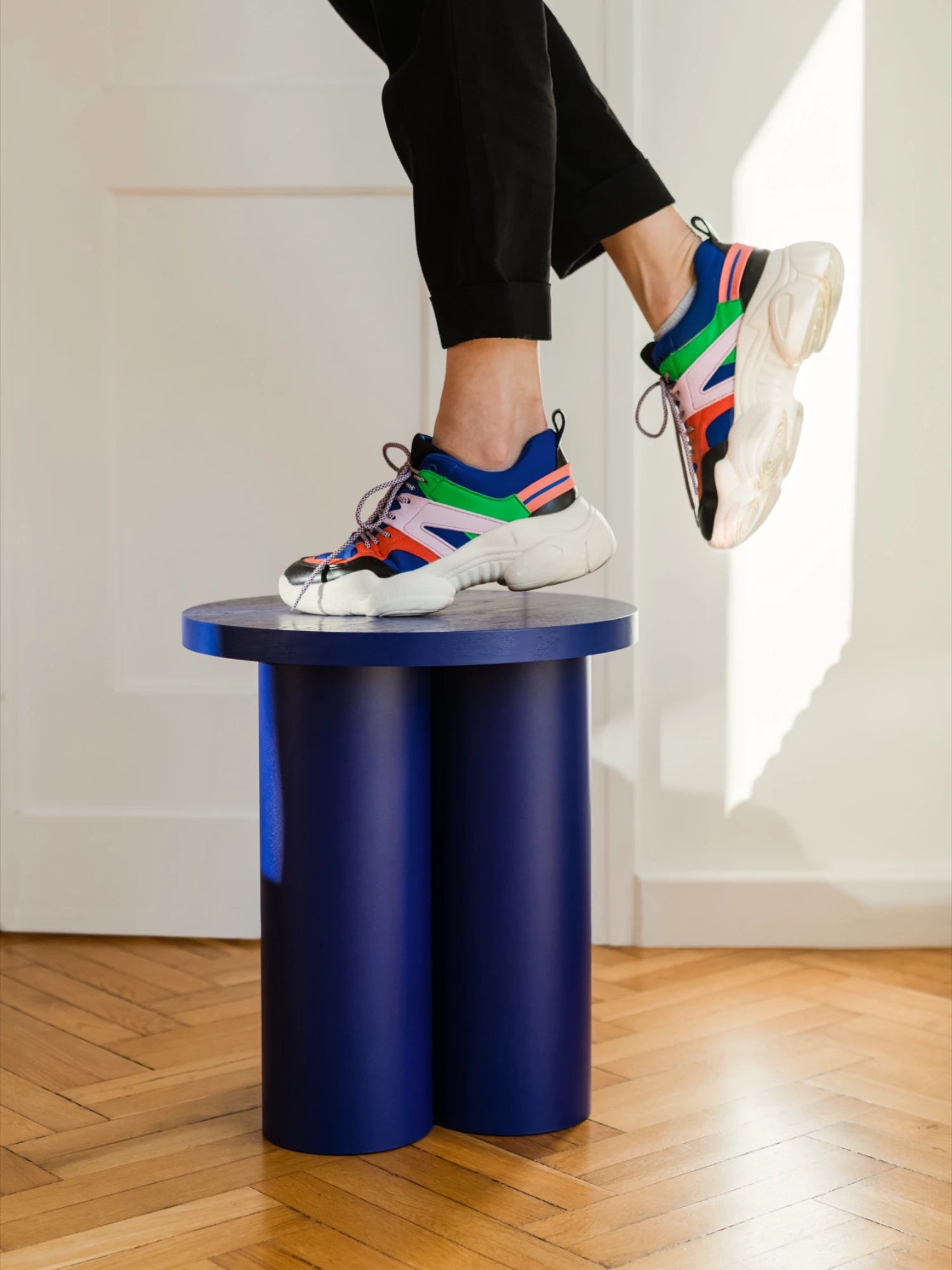 A person is shown standing with one foot on a dark blue Oly Stool. The stool has a round top and three thick, cylindrical legs. The person is wearing colorful sneakers and black pants, on a wooden floor. The other foot is in the air.