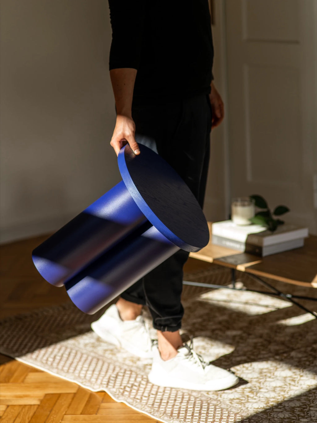 A person wearing a black t-shirt, black pants, and white sneakers is holding a dark blue Oly Stool. The stool has a round top and three thick, cylindrical legs. The person is standing on a textured rug over a wooden floor, with a wooden coffee table in the background.