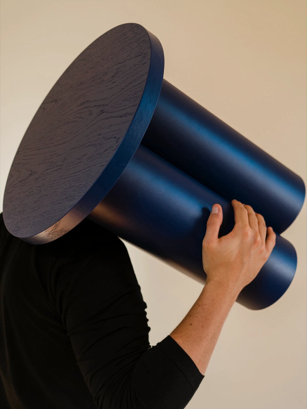 A person is shown from the back, holding a dark blue Oly Stool up to their shoulder. The stool has a round top and three thick, cylindrical legs. The wood grain is visible on the top of the stool.