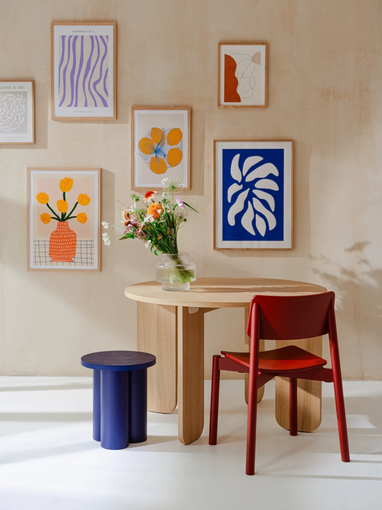 A small dining or cafe set is in a room with a light beige wall covered in several framed, colorful art prints. The set includes a round wooden table, a dark red chair, and a dark blue Oly Stool with a round top and three thick, cylindrical legs. A glass vase with a bouquet of flowers is on the table.
