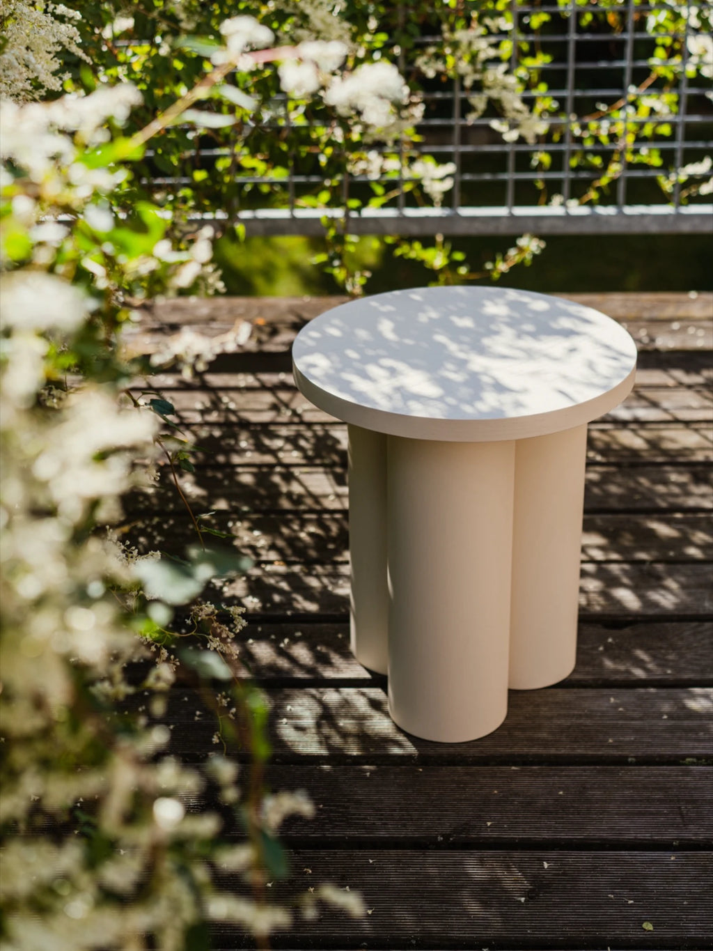 A modern, minimalist Oly Stool in a light Piazza Beige color is on a wooden deck. The stool has a round top and three thick, cylindrical legs that resemble pipes. It's placed near a railing, with white flowers and green foliage in the background.