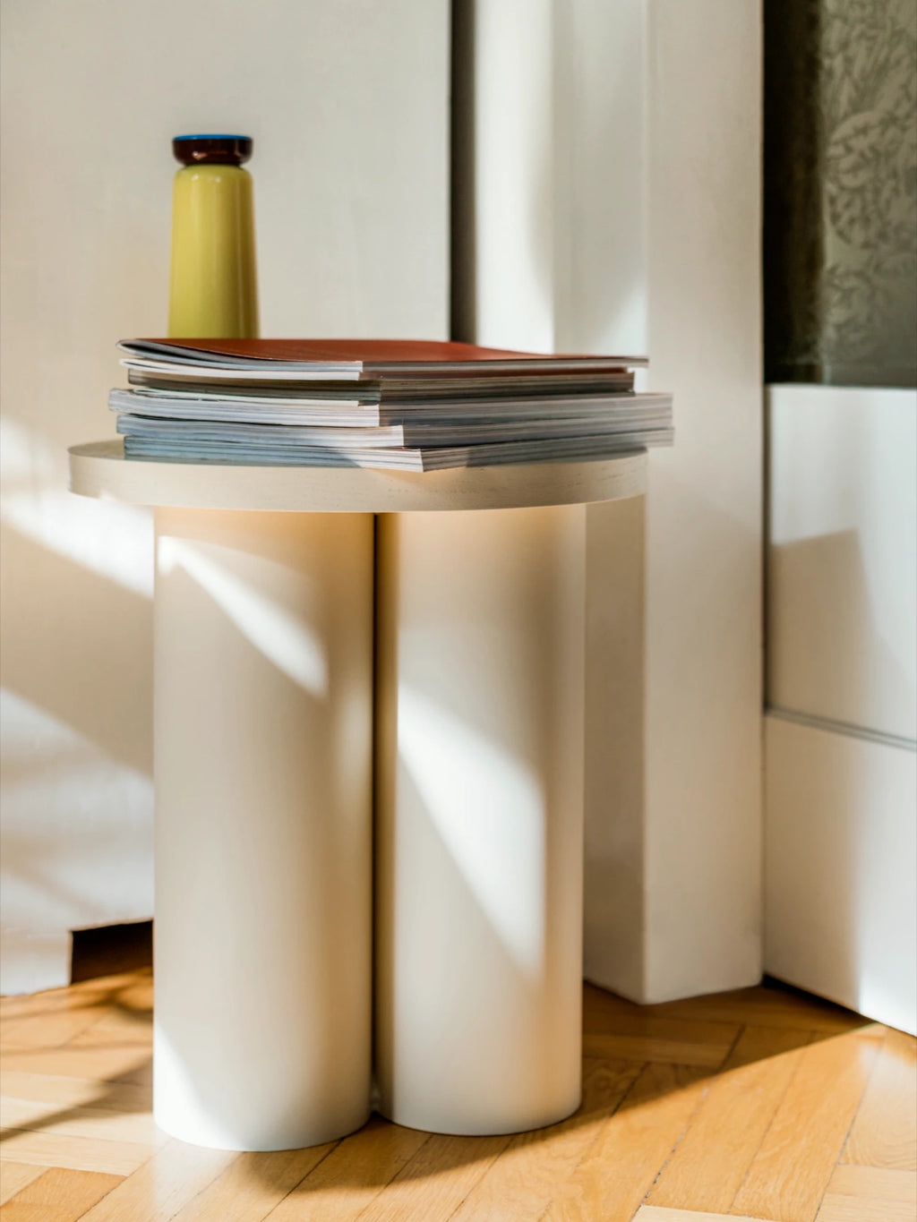 A light Piazza Beige Oly Stool is used as a side table indoors. It has a round top and three thick, cylindrical legs. A stack of magazines and a tall yellow bottle or vase are on the top. The stool sits on a wooden floor in a sunlit corner.