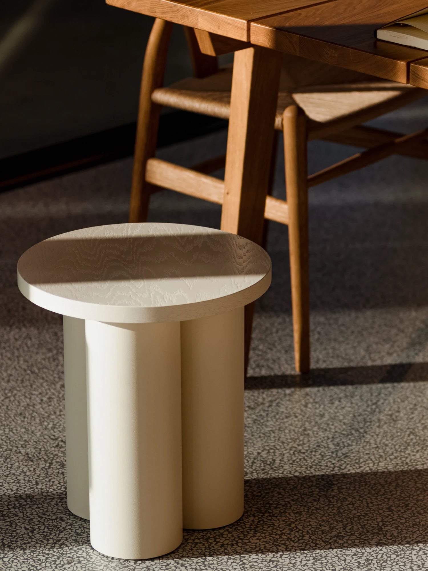 A small Oly Stool in a light Piazza Beige finish is on a gray speckled floor. The stool has a round top and three thick, cylindrical legs. Part of a wooden dining table and a matching chair are visible in the background, with sunlight casting shadows on the floor.