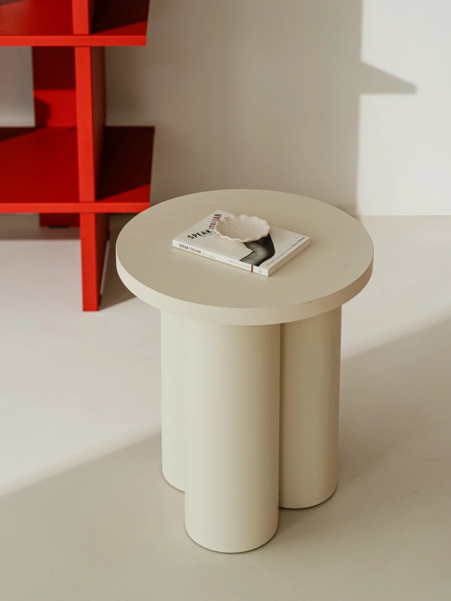 A light Piazza Beige Oly Stool with a round top and three thick, cylindrical legs is on a light-colored floor. The stool is used as a side table and is holding an open book with a decorative bowl on top. A red bookshelf is visible in the background.