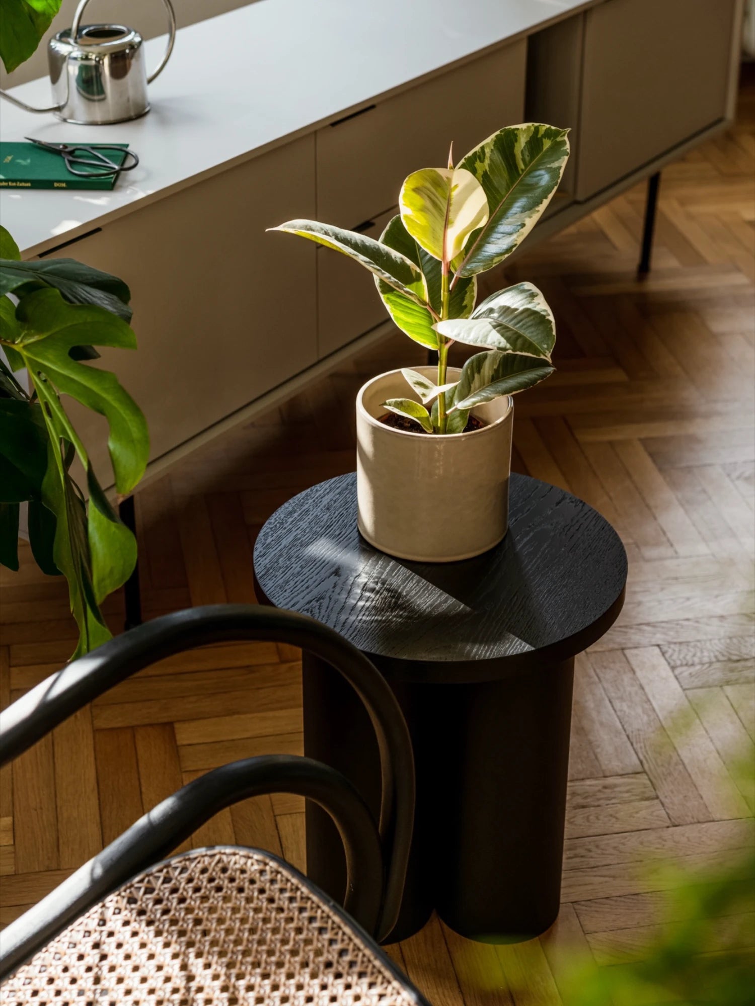 A black Oly Stool with a round top and three thick, cylindrical legs is used as a side table. It holds a potted plant with large variegated leaves. A white cabinet is in the background, and a chair with a cane back is in the foreground.