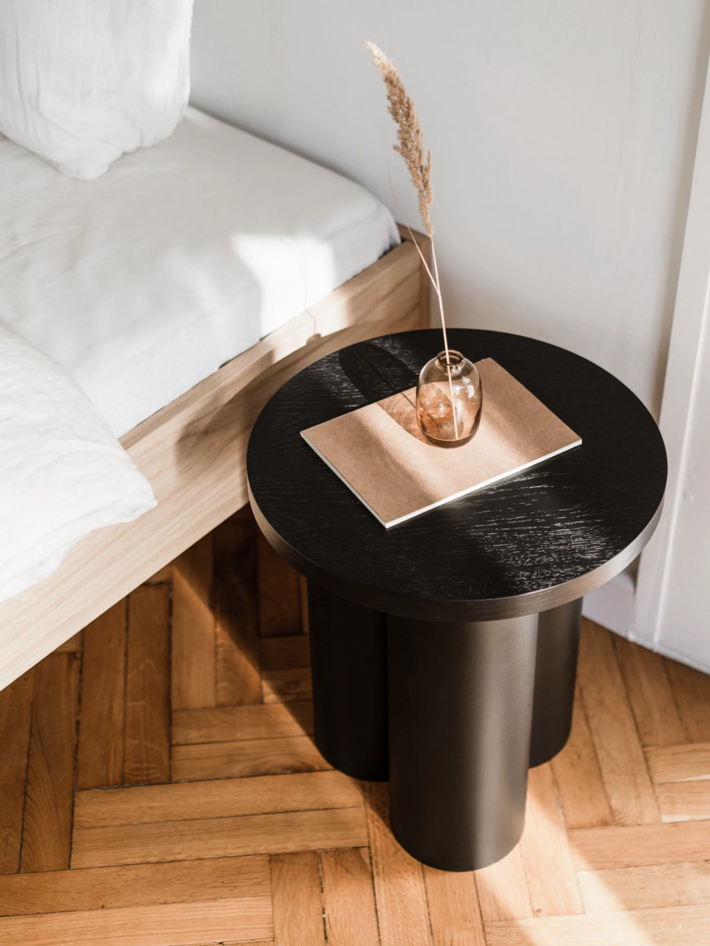 A round, matte black Oly Stool is used as a nightstand next to a light wood bed with white bedding. The stool has a round top and three thick, cylindrical legs. A small vase with a single dried plant stalk is on top, resting on a small, brown notebook. Sunlight casts a shadow across the scene.