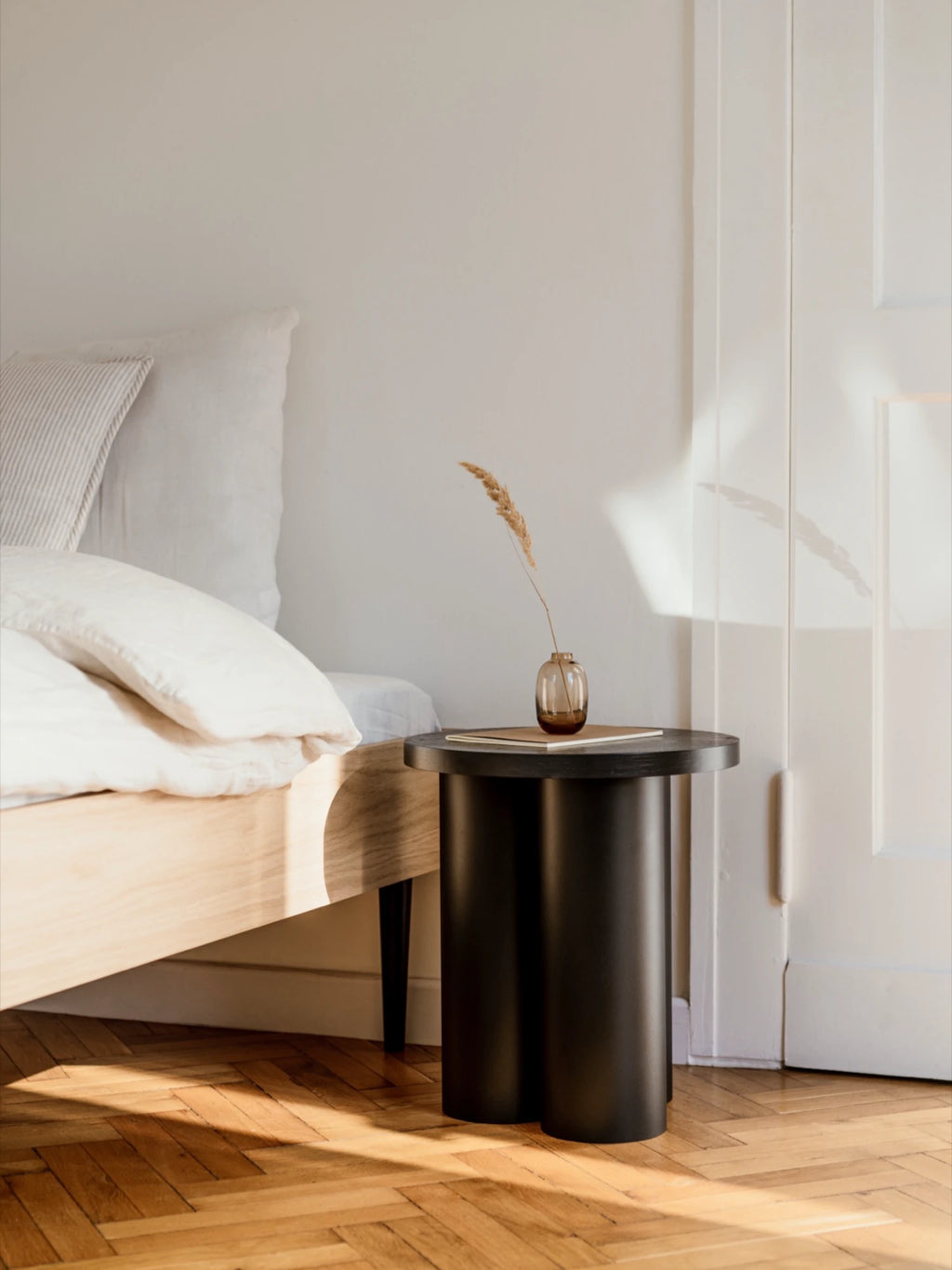 A minimalist Oly Stool in a matte black finish is used as a nightstand. The stool has a round top and three thick, cylindrical legs. A small brown vase with a single dried plant stalk is on top, next to a stack of books. The stool is next to a light wood bed with white bedding.
