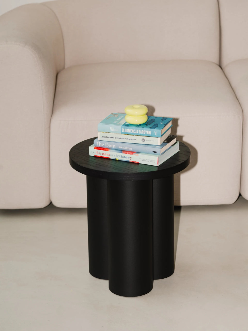 A round, matte black Oly Stool is used as a side table next to a light beige modular sofa. The stool has three thick, cylindrical legs and is holding a stack of books and a yellow decorative object.