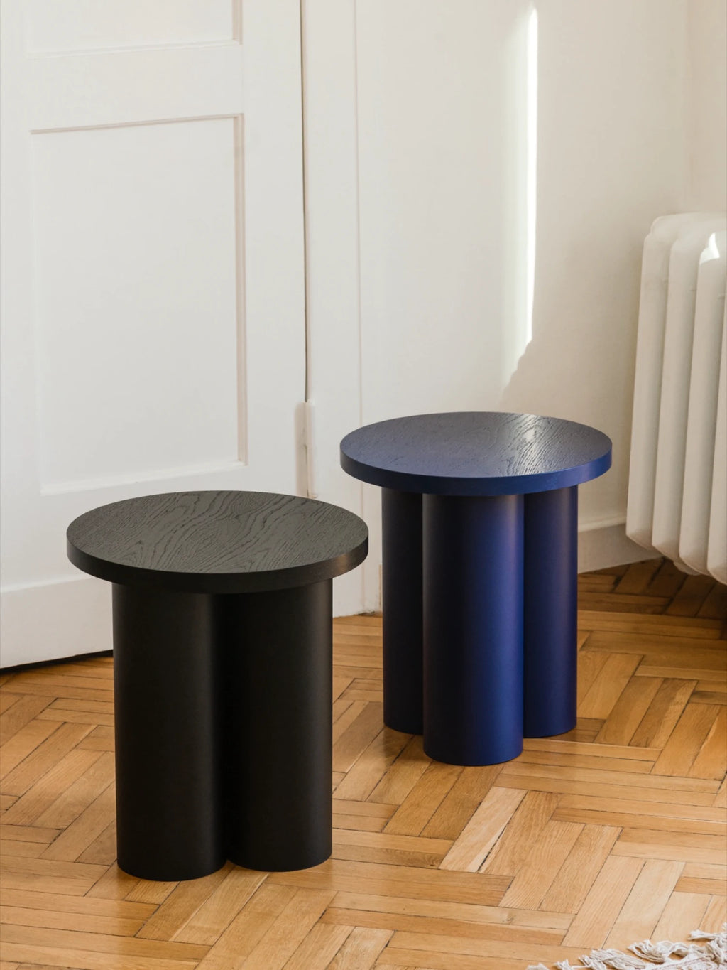 Two Oly Stools with round tops and three thick, cylindrical legs are on a wooden floor. The stools are different colors: one is a dark blue, and the other is a matte black. A white door and a radiator are visible in the background.