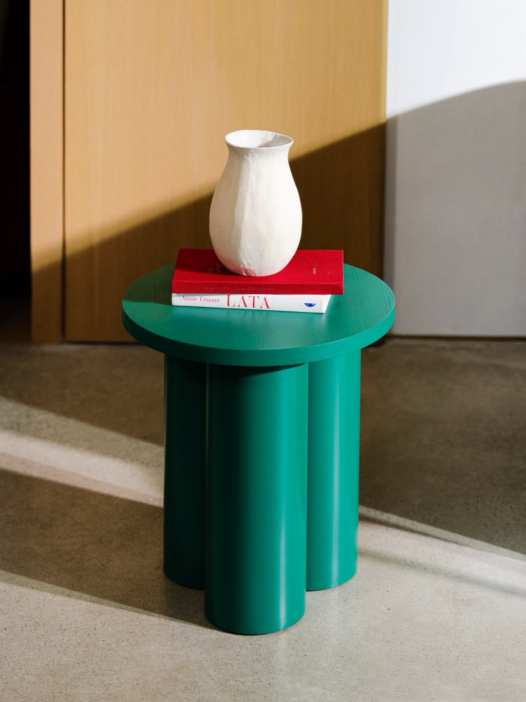 A dark green Oly Stool is used as a side table. It has a round top and three thick, cylindrical legs. A red and a green book are stacked on top, with a textured white vase resting on the books. The stool is on a light-colored floor, with a wooden door partially visible in the background.