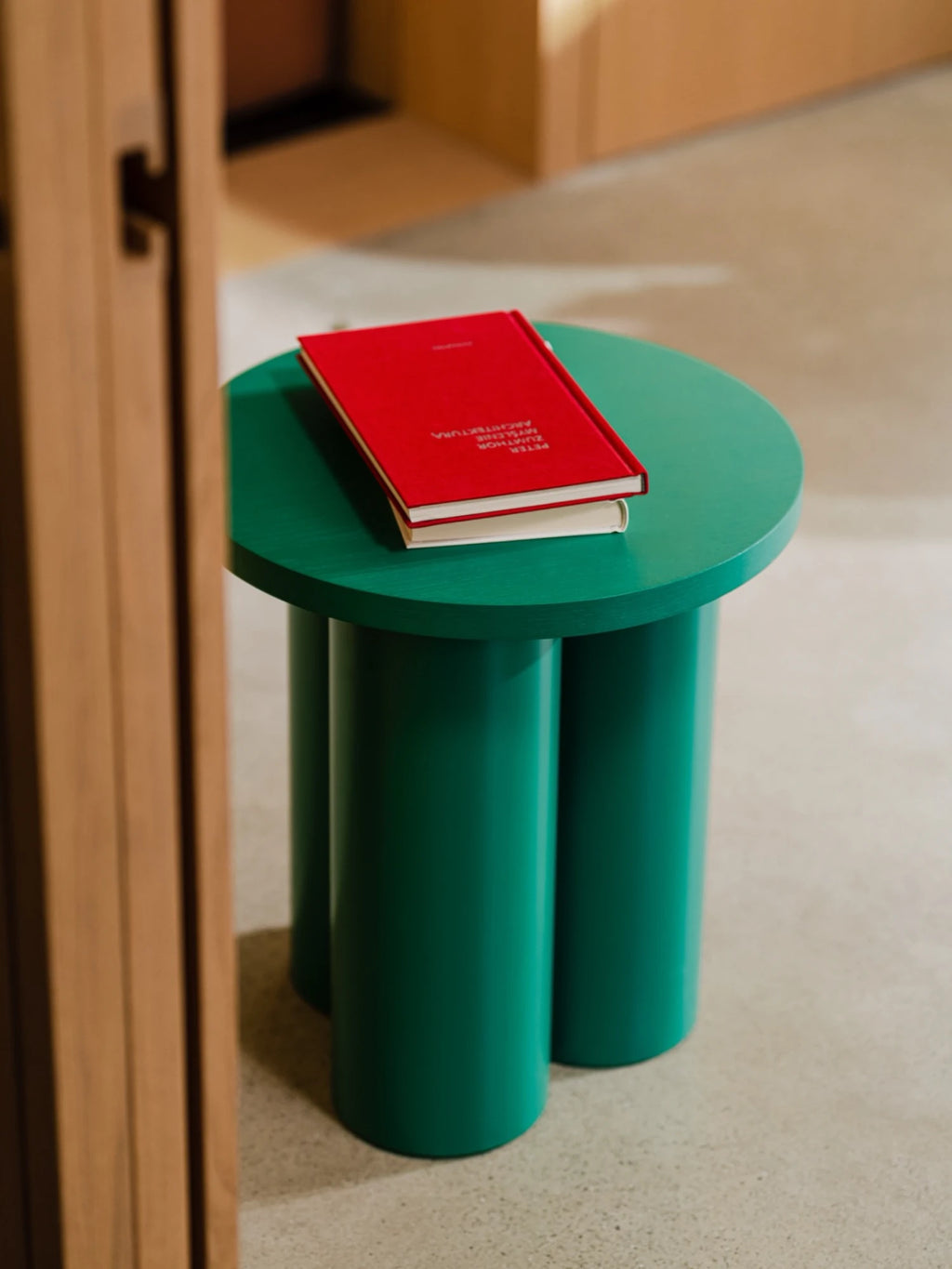 A vibrant Oly Stool in a dark green color is placed on a light-colored floor. The stool has a round top and three thick, cylindrical legs, with two books with a red cover stacked on top. A wooden door frame is visible on the left side of the image.