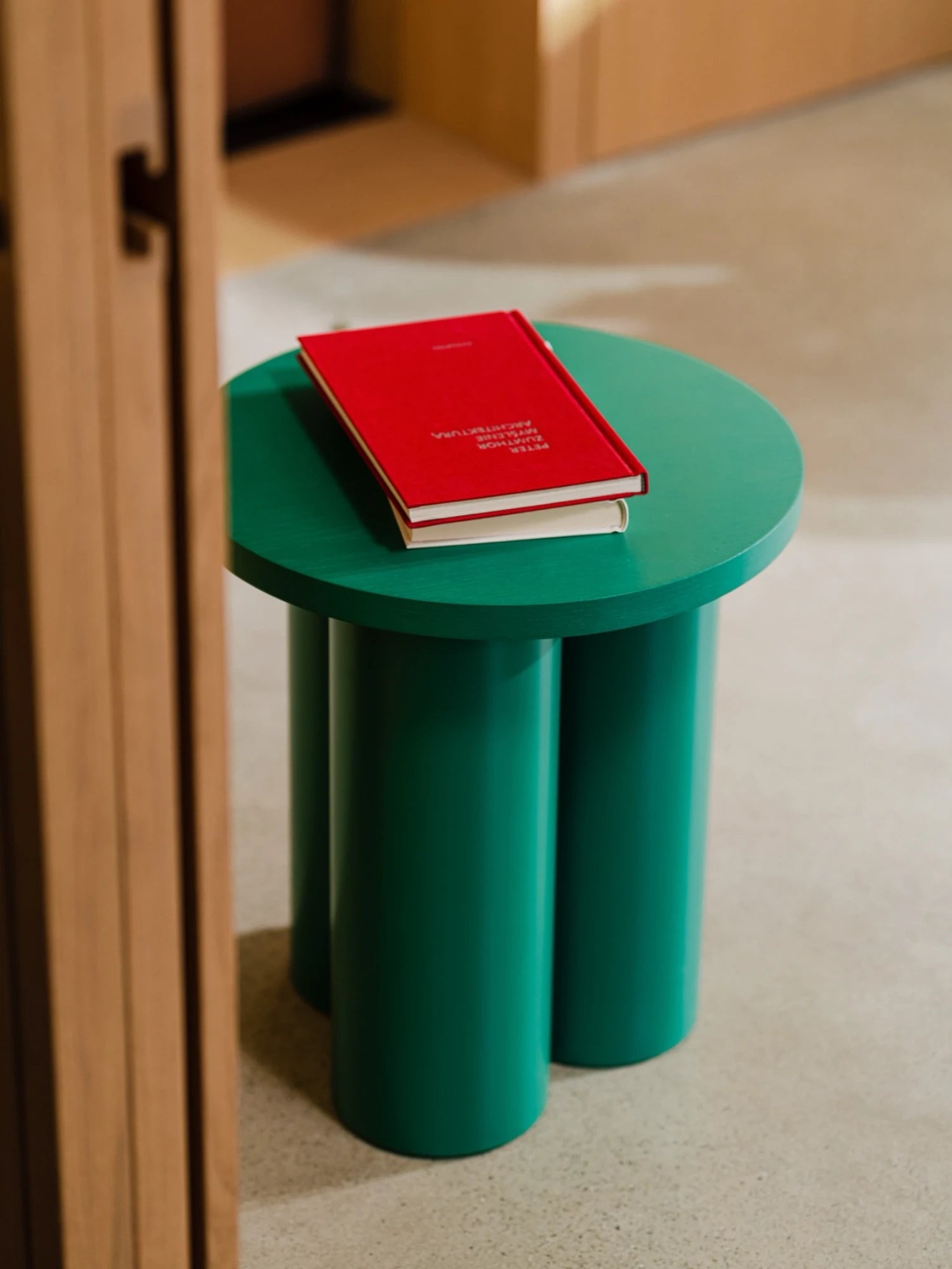 A vibrant Oly Stool in a dark green color is placed on a light-colored floor. The stool has a round top and three thick, cylindrical legs, with two books with a red cover stacked on top. A wooden door frame is visible on the left side of the image.