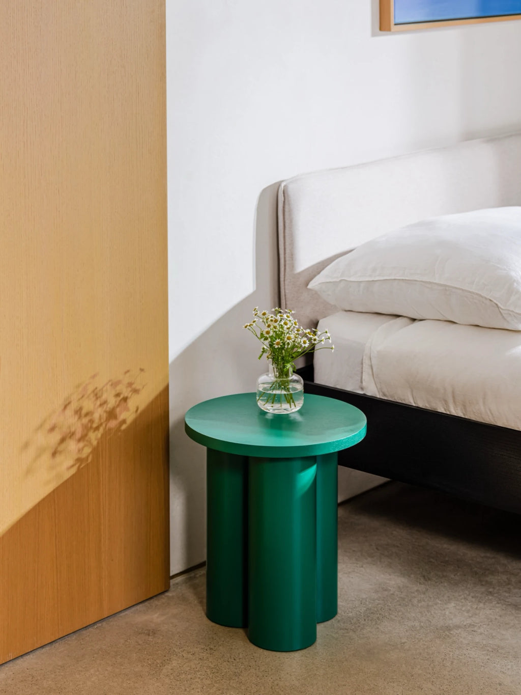 
A small Oly Stool in a dark green color is used as a nightstand. The stool has a round top and three thick, cylindrical legs, and it's holding a small glass vase with white flowers. The stool is next to a bed with a white headboard and pillows, with a wooden door visible on the left.