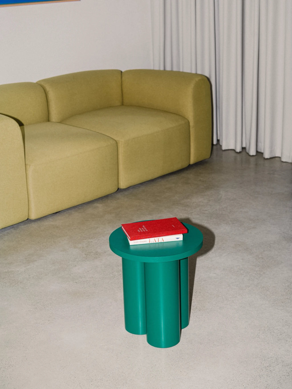 A vibrant Oly Stool in a dark green color is placed in a minimalist living room. The stool has a round top and three thick, cylindrical legs, and it's holding two books with a red cover on top. A light green modular sofa and a set of white curtains are in the background.