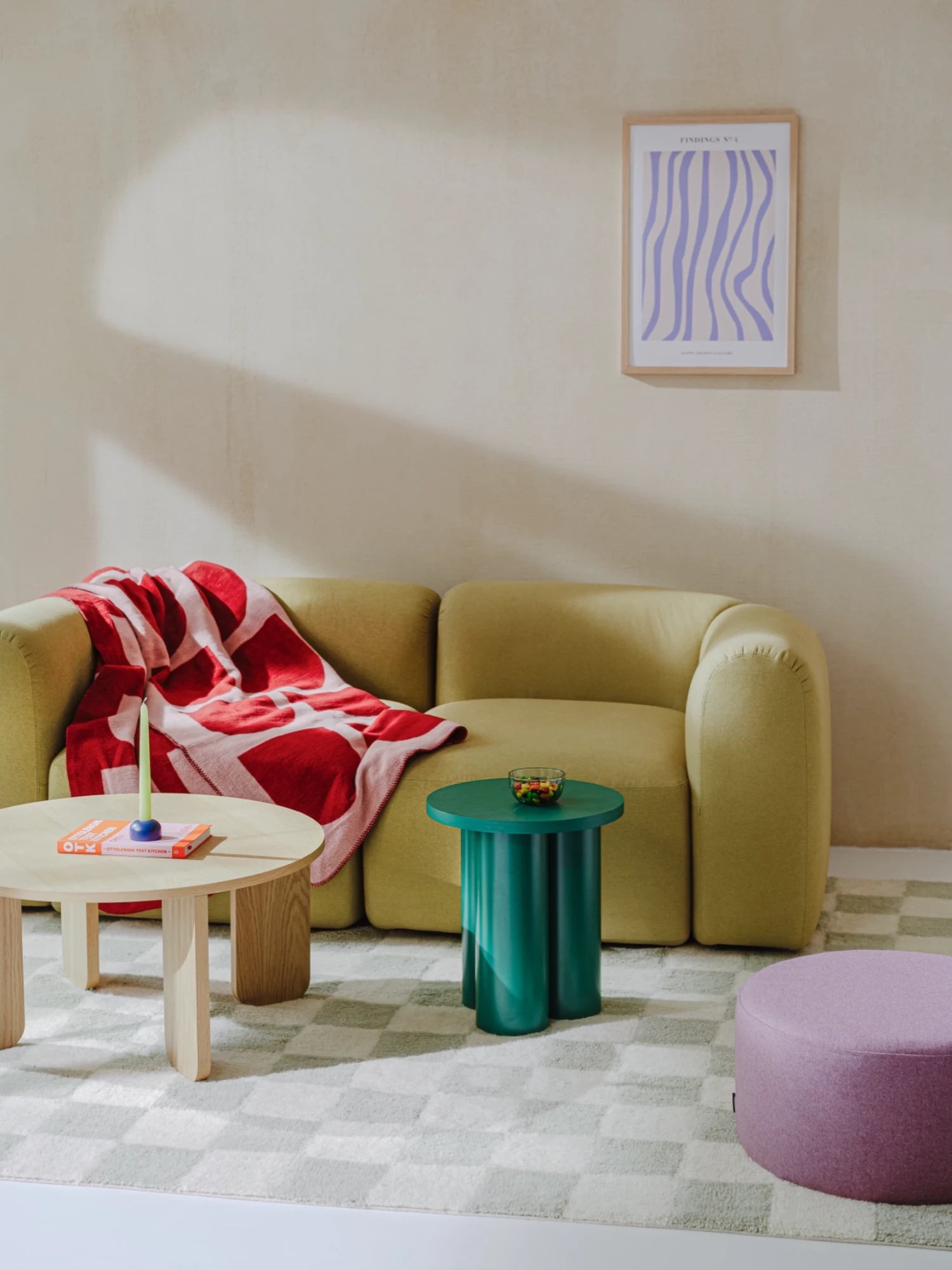 A vibrant Oly Stool in a dark green color is placed in a living room, holding a small bowl of candy. The stool has a round top and three thick, cylindrical legs. It is positioned next to a light green modular sofa and a round wooden coffee table. A colorful blanket is draped over the sofa, and a checked rug covers the floor.