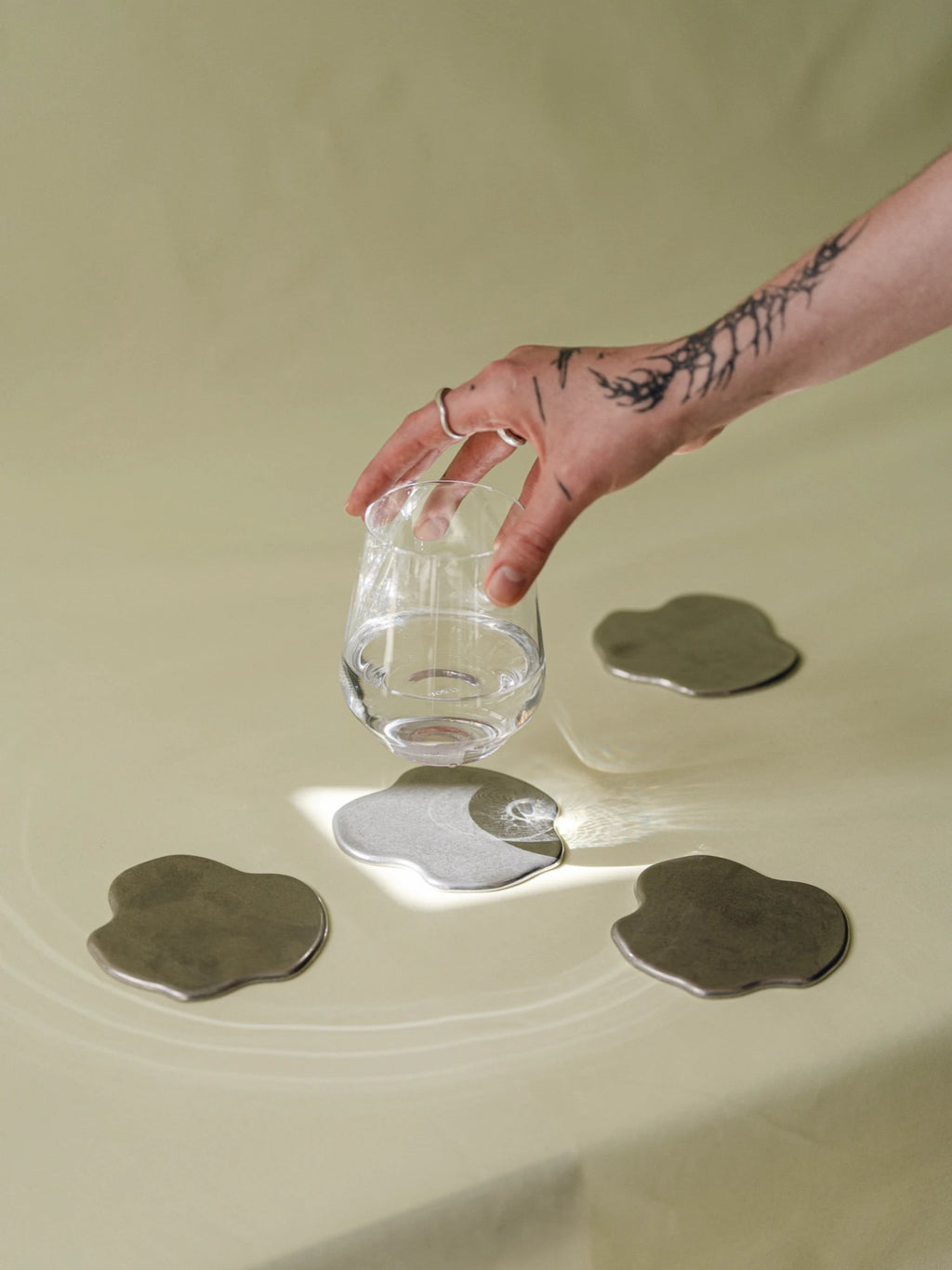 noo.ma Plama Stainless Steel Coaster - Set of 4, a collection of modern minimalist coasters with an organic, fluid, irregular shape. Crafted from durable stainless steel, some feature a brushed metallic grey finish while one boasts a polished, reflective silver surface. These contemporary metal coasters are perfect for protecting furniture in a modern living room, home office, or stylish dining area.