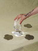 Hand holding a glass over water with metallic coasters on a beige surface