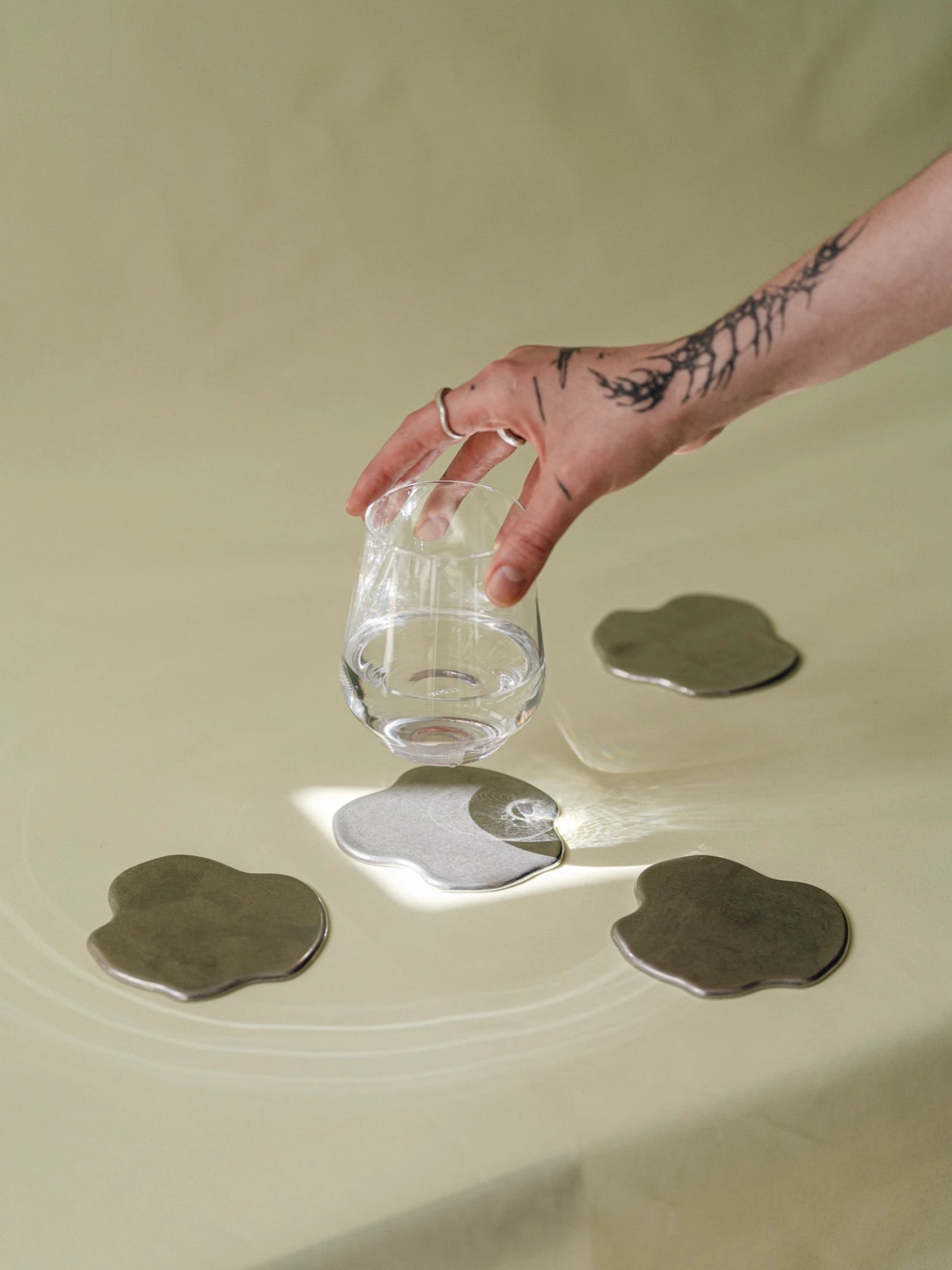 Hand holding a glass over water with metallic coasters on a beige surface