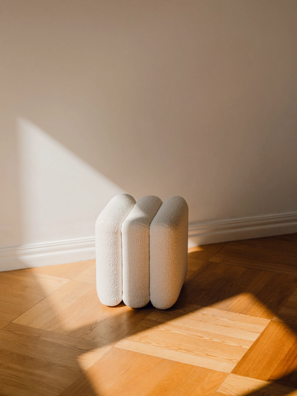 noo.ma Radd Pouf, a cream-colored organic modern pouf upholstered in soft textured bouclé fabric, featuring a unique sculptural shape with three rounded, pill-like segments and a clean contemporary aesthetic. This versatile accent stool is perfect for adding minimalist style and cozy texture to living rooms, bedrooms, or entryways.