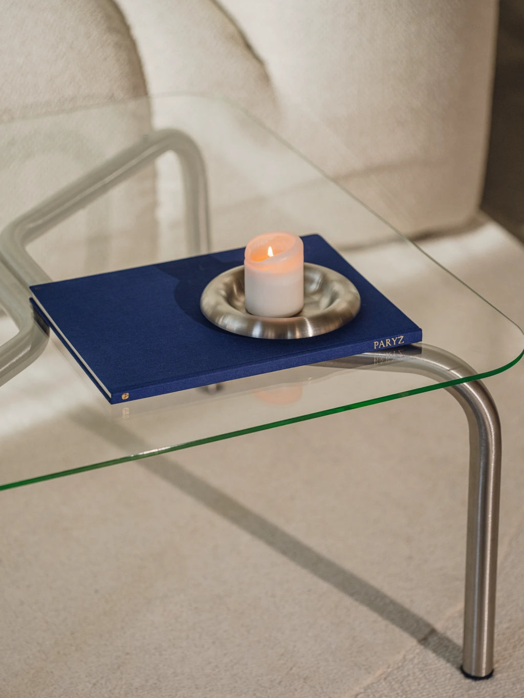 Glass side table with a blue tray holding a candle and decorative item on a light-colored floor.