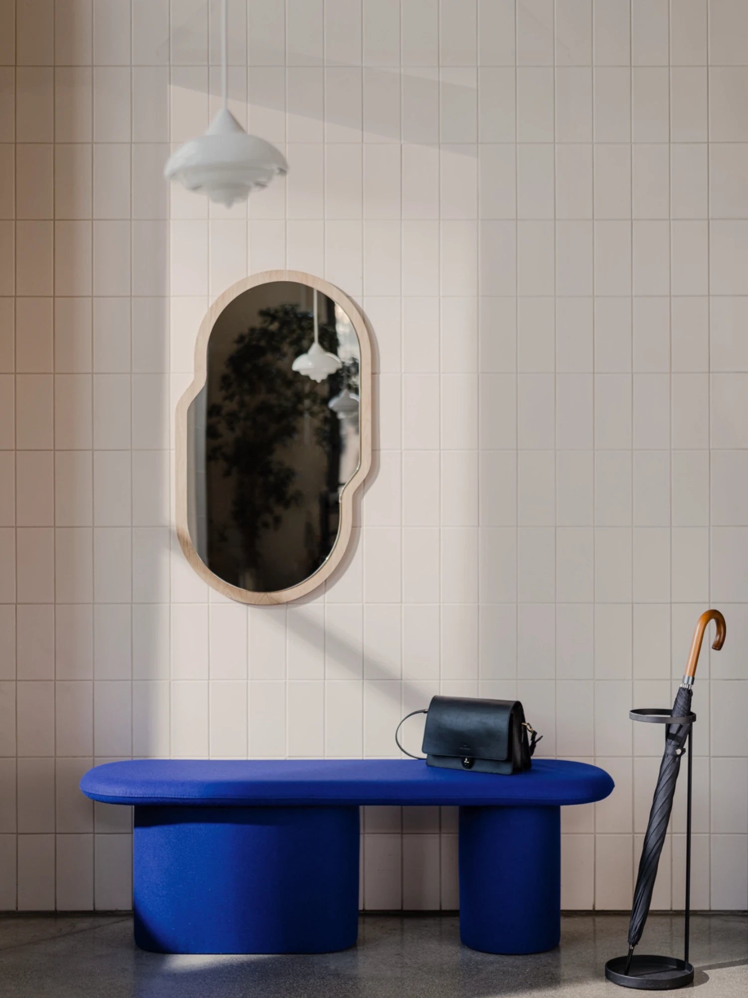 A low, pill-shaped upholstered bench in a deep blue color is in an entryway with white tiled walls. The bench has two thick, cylindrical legs and a black bag is resting on it. A unique, irregularly shaped mirror with a light wood frame is hanging on the wall above the bench. An umbrella stand with a black umbrella is on the right side of the image.