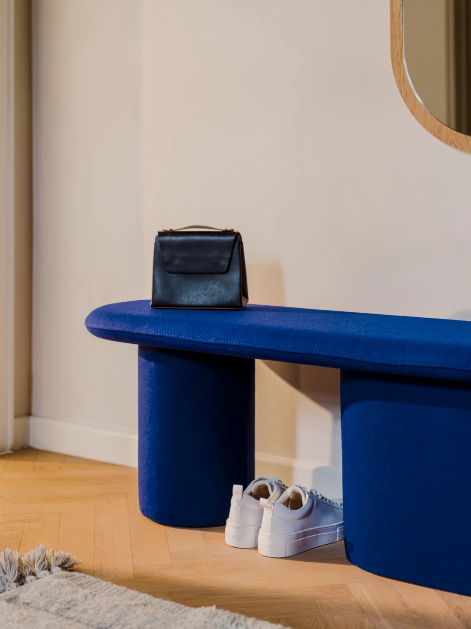 A low, upholstered, pill-shaped bench in a deep blue color is on a light wooden floor. The bench has two thick, cylindrical legs. A black handbag is resting on the bench, and a pair of white sneakers is on the floor beneath it.