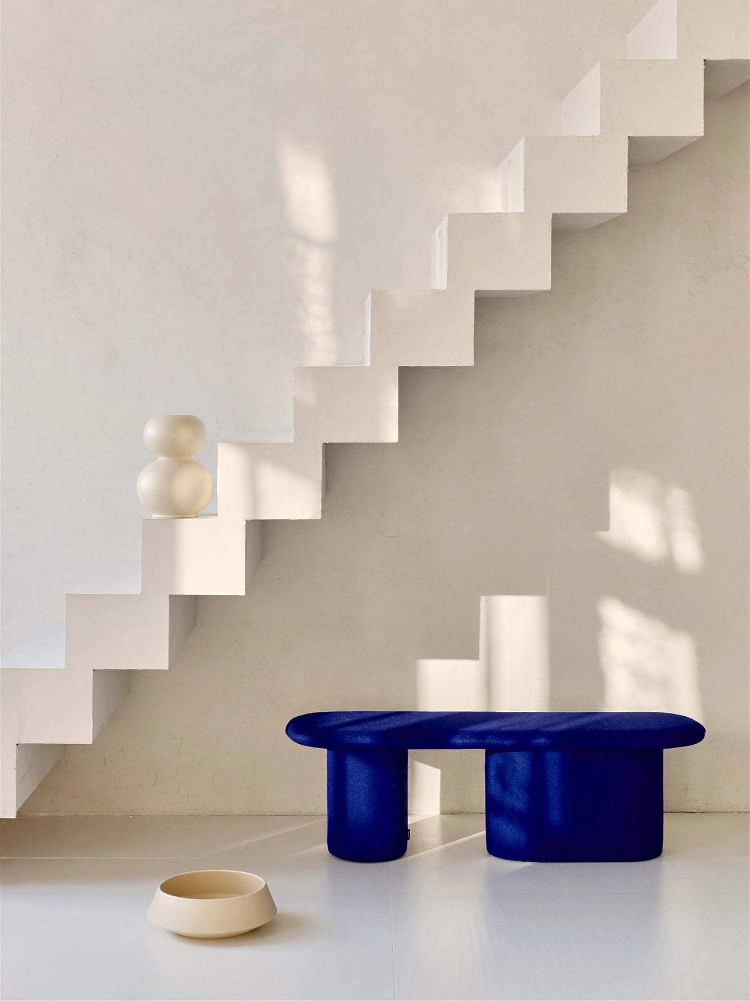 noo.ma Rull Bench, a striking deep royal blue upholstered bench featuring a plush velvet texture and a distinctive organic, sculptural design. It showcases a long, rounded top surface supported by two robust, cylindrical bases, creating a bold, contemporary aesthetic. Ideal as a statement seating piece for a minimalist living room, modern entryway, or a vibrant bedroom accent.