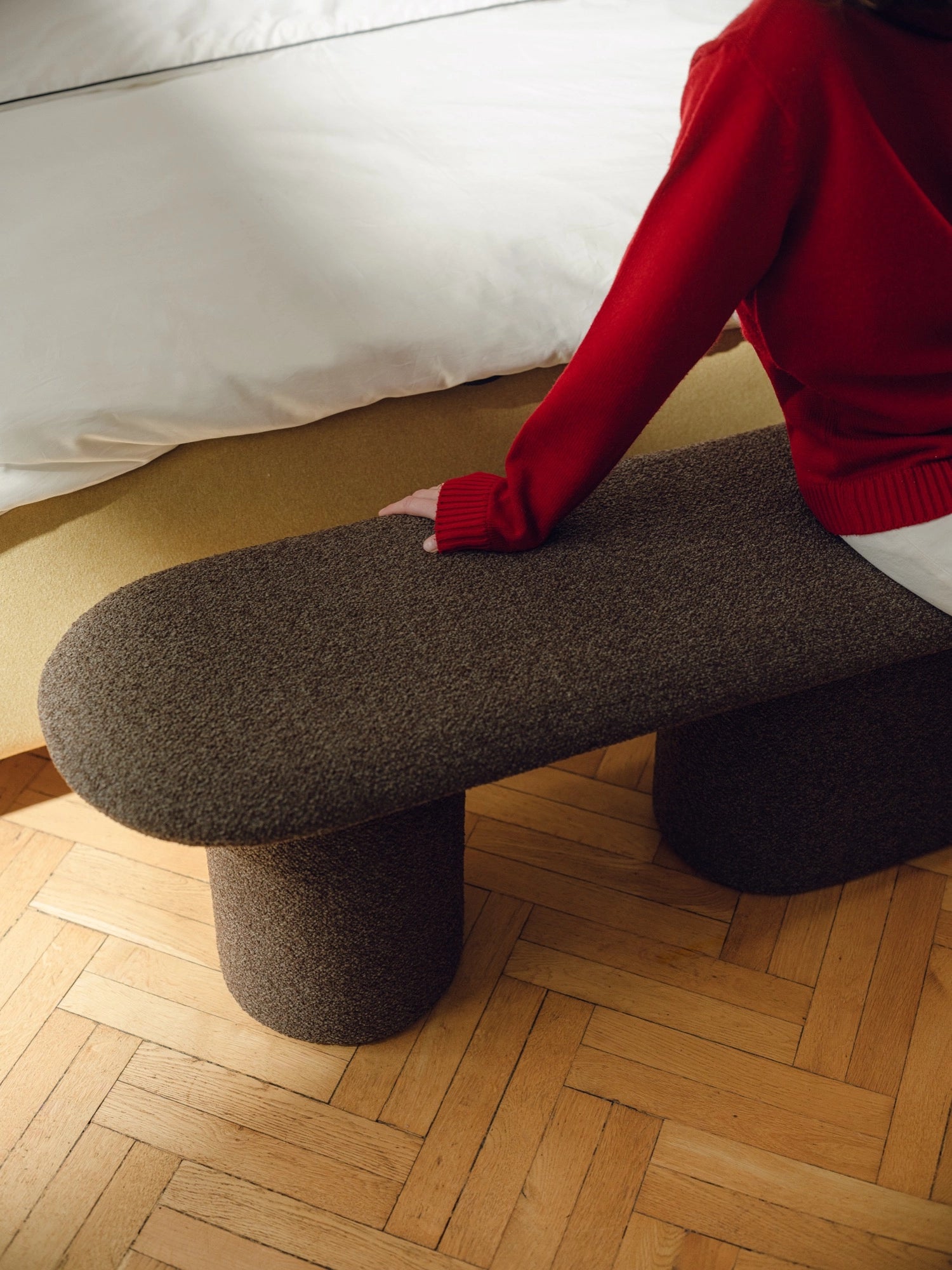 noo.ma Rull Bench, a dark brown upholstered bench featuring an elongated oval top and two sturdy cylindrical legs, all meticulously covered in a rich, nubby bouclé-like textured fabric. This organic modern and minimalist sculptural bench offers stylish and comfortable seating, ideal for bedroom entryways, living room accents, or contemporary hallways.