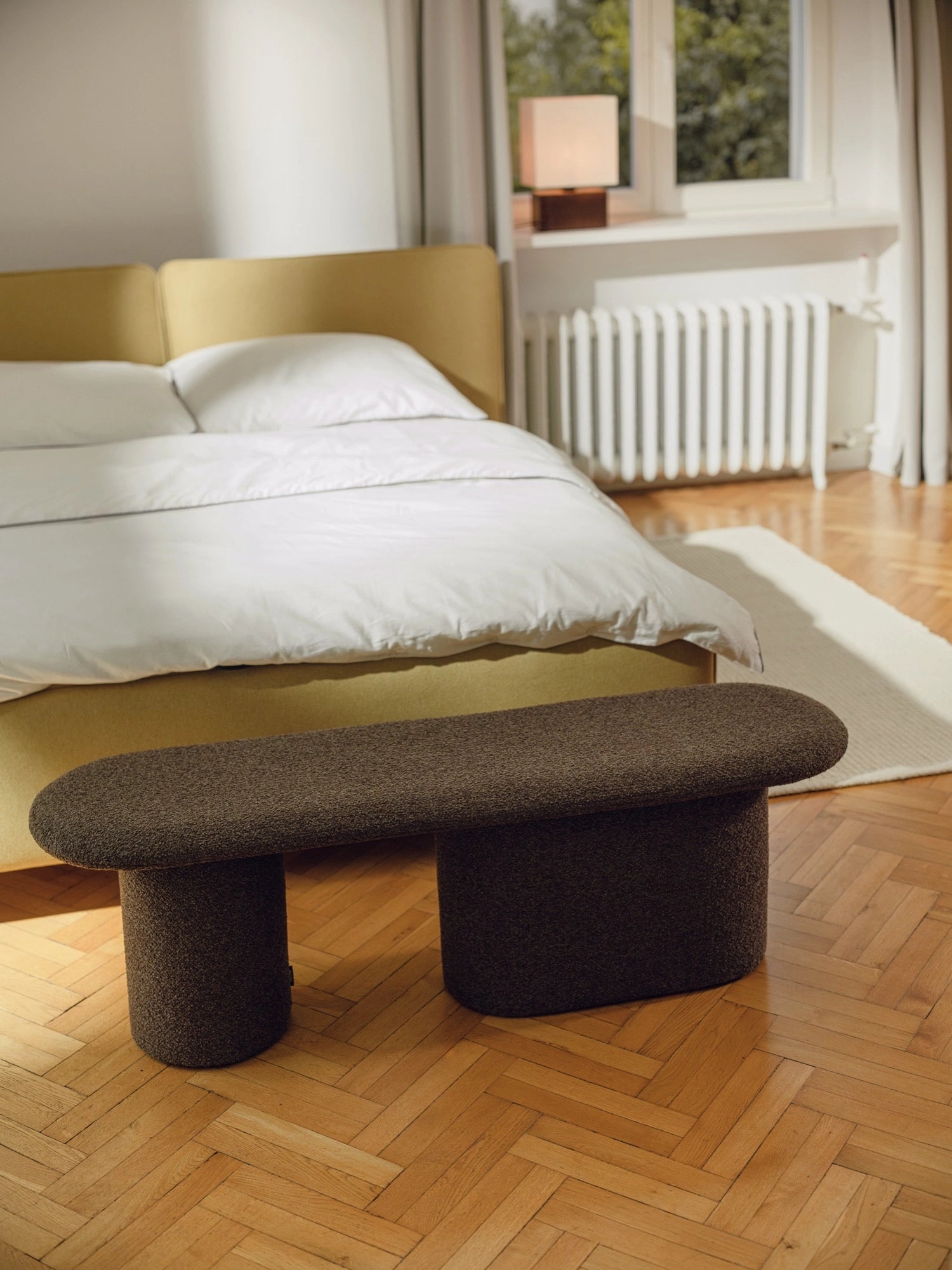 noo.ma Rull Bench, a dark brown bouclé upholstered bench featuring a soft, textured fabric and an organic, pill-shaped top with contrasting cylindrical and rectangular block legs. This sculptural modern bench is perfect as an end-of-bed bench for bedrooms or as minimalist seating in contemporary living rooms and entryways.