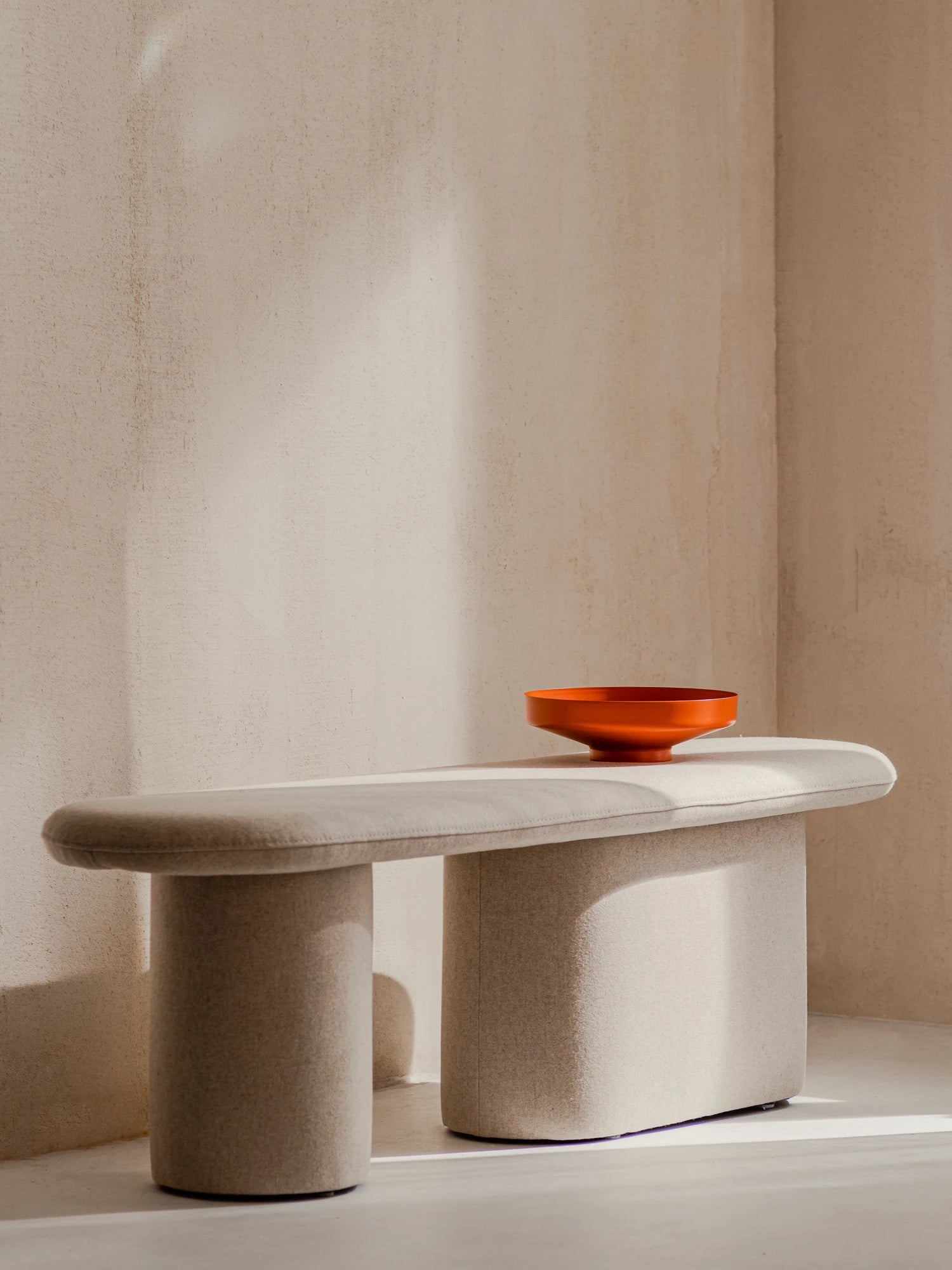 A long, pill-shaped upholstered bench in a classic beige color is on a light floor in a sunlit corner. The Rull Bench has two different legs: one is a thick cylindrical shape, and the other is a thicker, rectangular-like shape. An orange decorative bowl is on top of the bench.