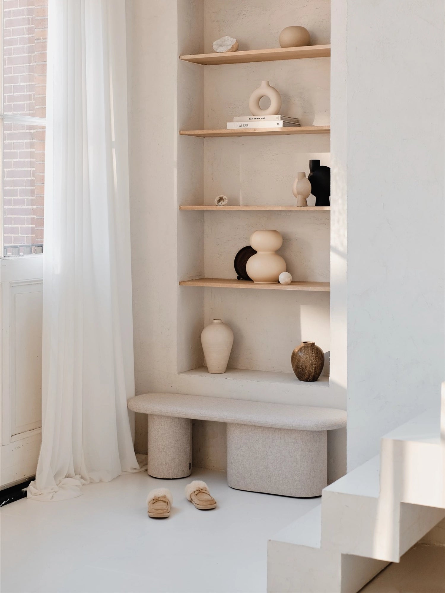 noo.ma Rull Bench, an organic modern upholstered bench featuring a softly curved top and two distinct, rounded bases, all in a light beige textured fabric. This minimalist and contemporary seating piece offers versatile accent styling for an entryway, living room, or bedroom.