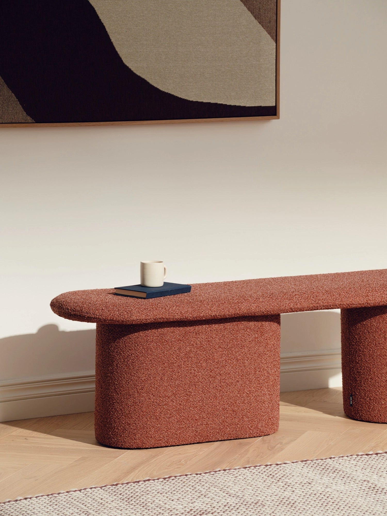 noo.ma Rull Bench, a contemporary organic modern bench upholstered in a rich burnt orange textured bouclé-like fabric, featuring a soft, elongated oval top and two rounded, cylindrical bases. This minimalist curved bench offers cozy seating or a stylish surface for living rooms, entryways, or bedroom accents.