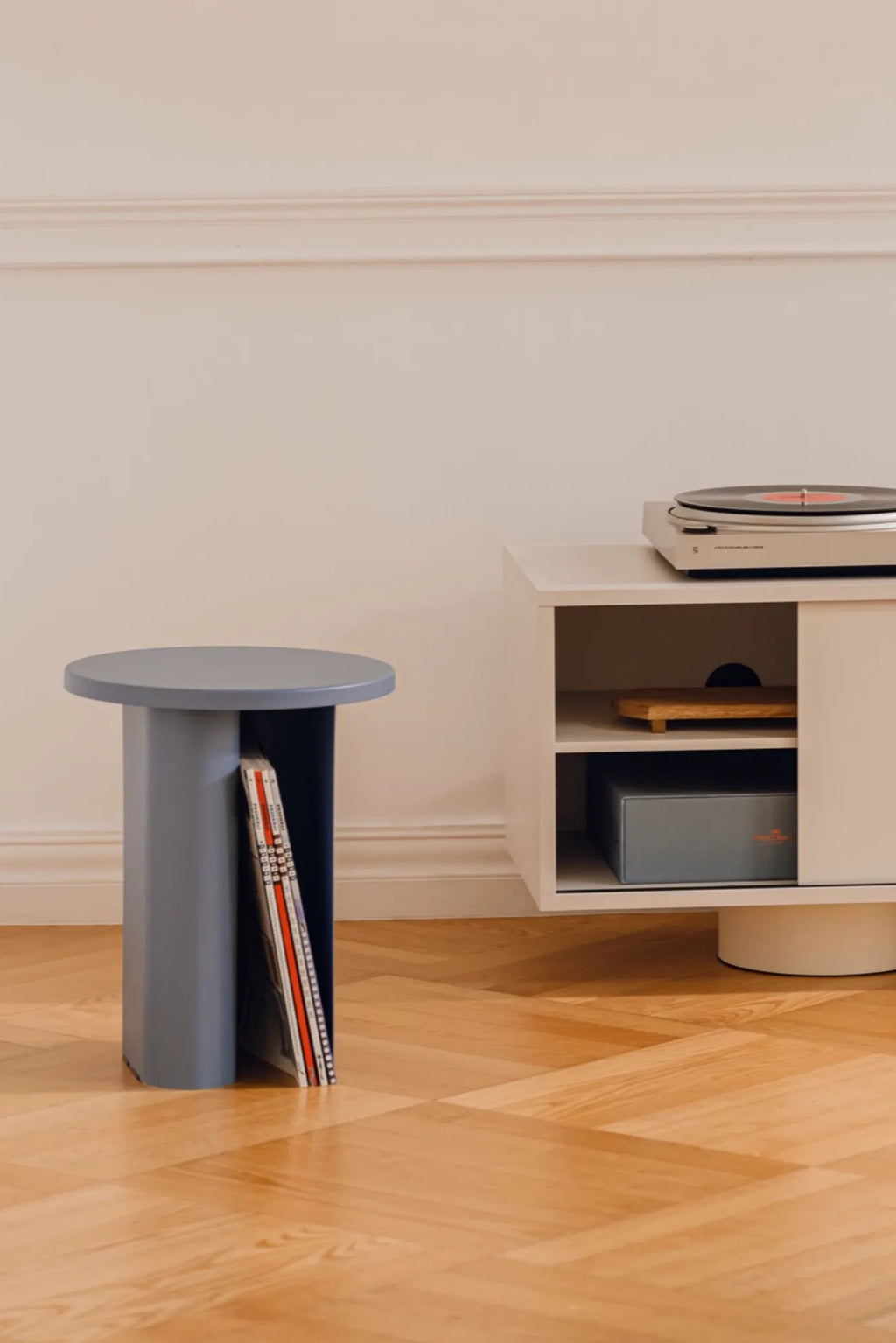 noo.ma Sato Side Table, a modern minimalist side table crafted from painted wood with a dusty blue matte finish, featuring a round top and a sculptural cylindrical base. Its unique design includes a dark navy blue vertical slot, perfect for stylishly storing vinyl records, magazines, or books, blending contemporary aesthetics with functional storage. This versatile accent table is ideal for Scandinavian living rooms, modern bedrooms as a nightstand, or creative home offices.