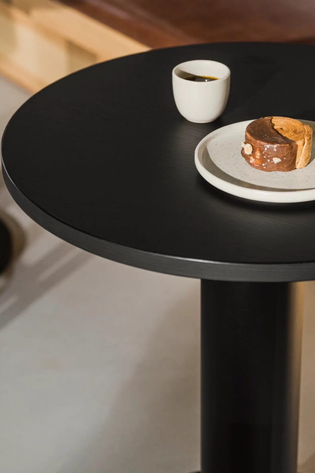 noo.ma Sool Café Table, a modern minimalist round dining table crafted from matte black solid wood, featuring a smooth circular tabletop with subtle wood grain texture and a sleek cylindrical pedestal base. Ideal for compact modern kitchens, small dining rooms, or contemporary café and bistro settings.