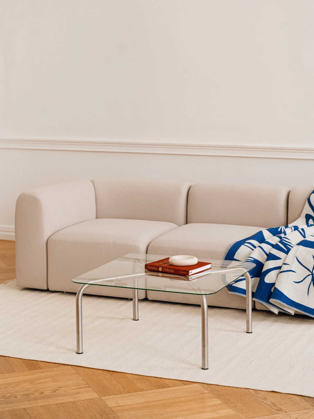 noo.ma Uku Coffee Table, a rectangular glass coffee table with brushed silver legs, positioned in a contemporary living room. A light beige modular sofa with a blue and white patterned throw blanket and a red book on the table complete the inviting scene.