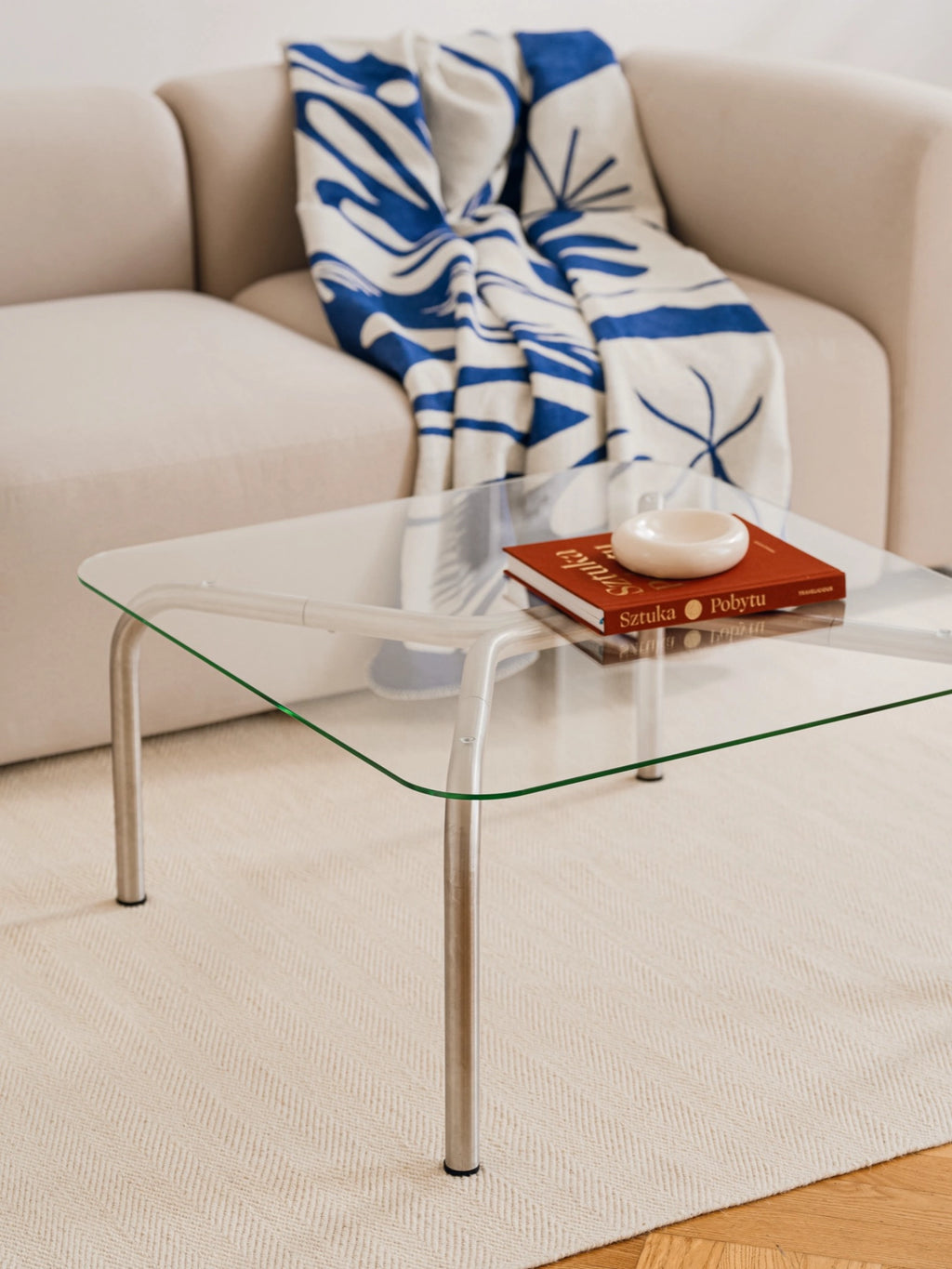 noo.ma Uku Coffee Table, a minimalist square glass coffee table with sleek silver metal legs, holding a book and an ashtray, positioned on a textured cream rug beside a beige sofa.