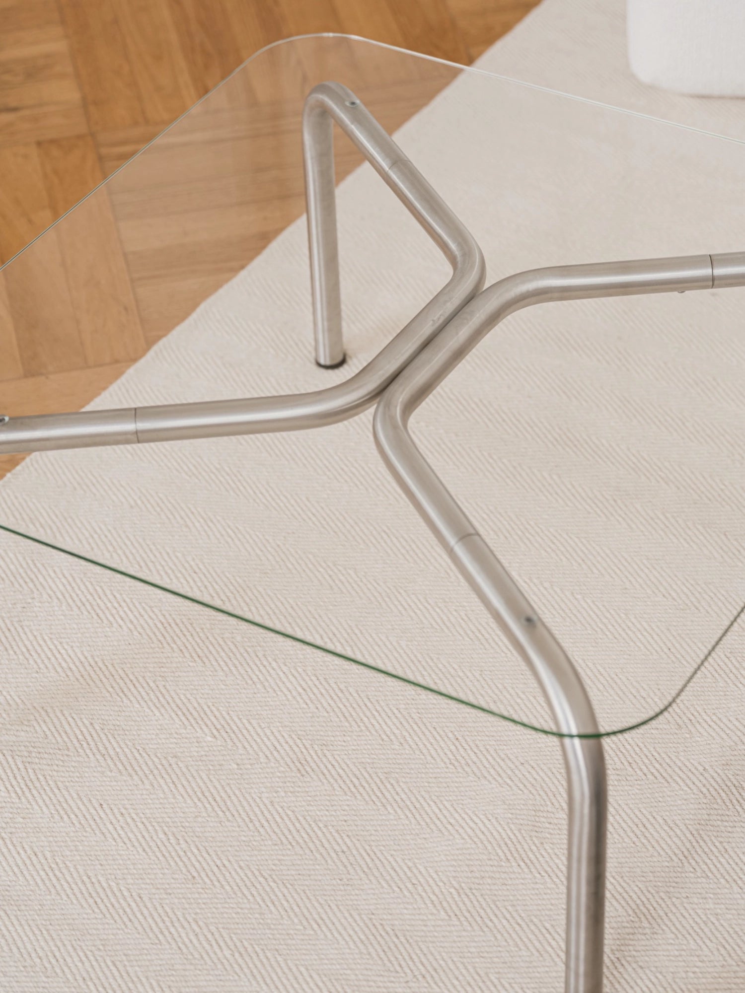 noo.ma Uku Coffee Table, showcasing the intricate silver metal base structure with curved legs and cross-bracing beneath the clear glass top. The table rests on a textured cream rug, with a warm wooden floor visible in the background.