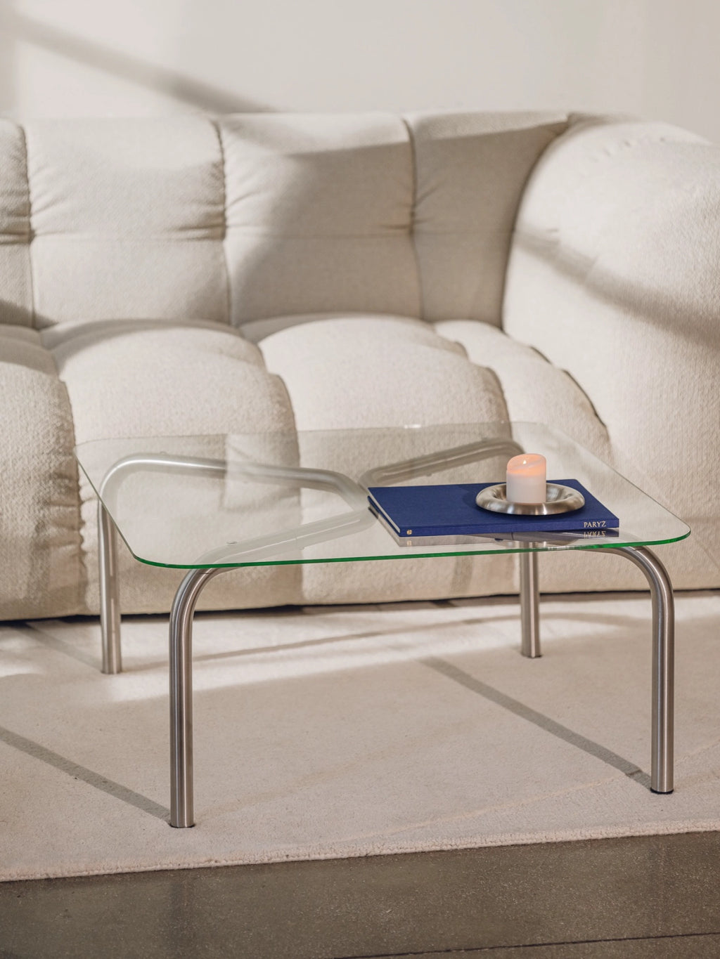 noo.ma Uku Coffee Table, a modern square glass top coffee table with sleek silver metal legs, featuring a minimalist design. Styled with a blue book and lit candle, placed on a light textured rug next to a cream bouclé sofa.