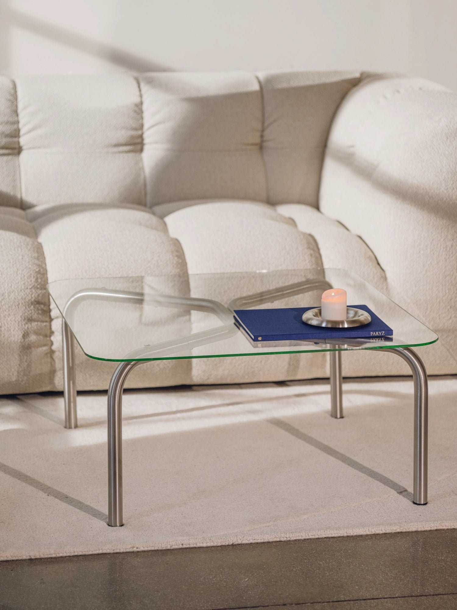noo.ma Uku Coffee Table, a modern square glass top coffee table with sleek silver metal legs, featuring a minimalist design. Styled with a blue book and lit candle, placed on a light textured rug next to a cream bouclé sofa.