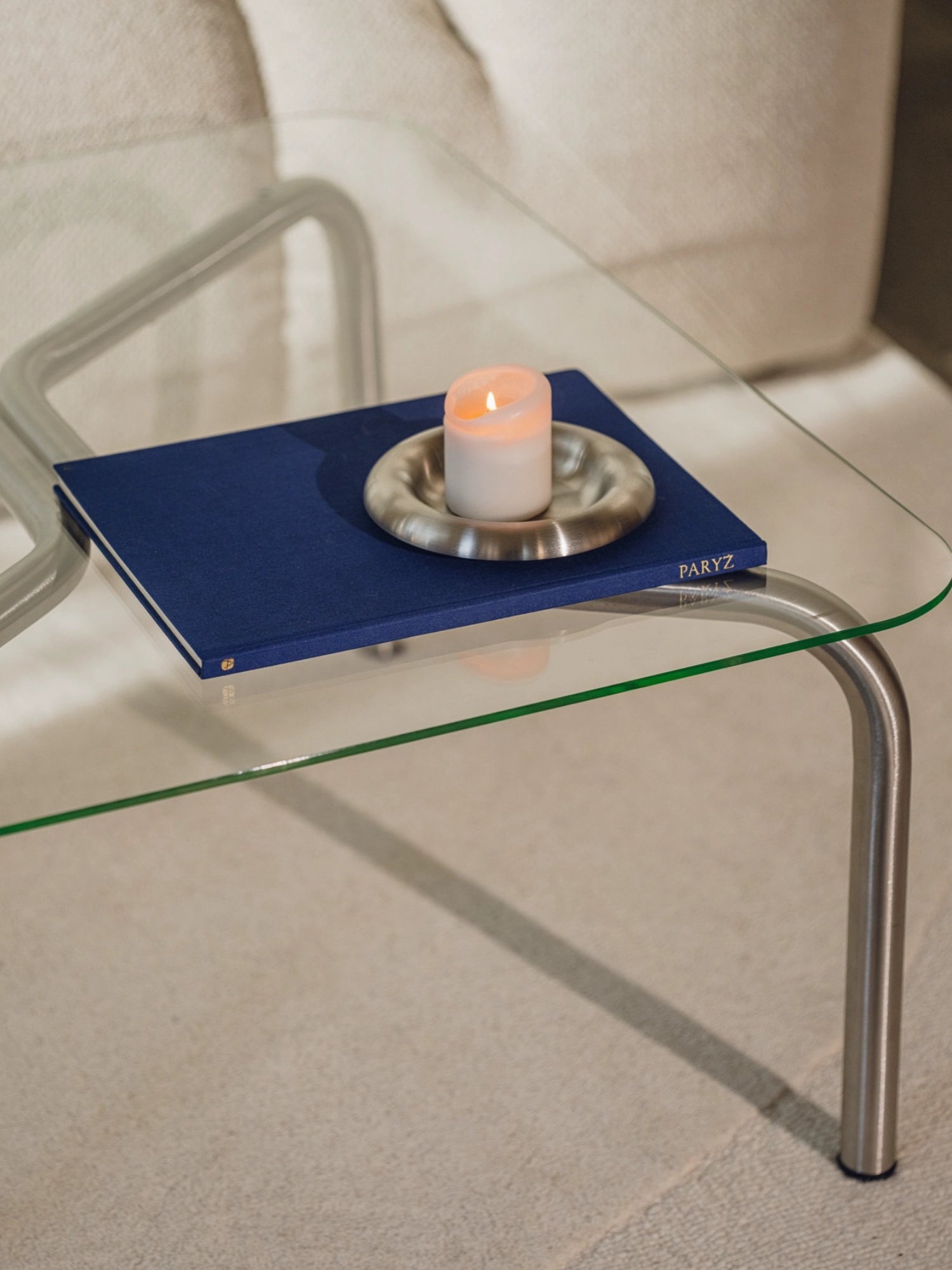 noo.ma Uku Coffee Table, a close-up view of the square glass top and one curved silver metal leg. A dark blue book and a lit candle in a silver holder are elegantly placed on the transparent surface, highlighting the table's modern aesthetic.