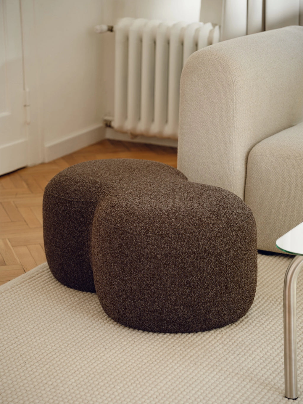 noo.ma Umbo Pouf, a dark brown organic modern pouf crafted from soft, textured bouclé fabric, featuring a distinctive rounded, connected silhouette resembling a figure-eight. This inviting contemporary ottoman serves as versatile accent seating or a stylish footrest for minimalist living rooms and cozy bedroom lounges.