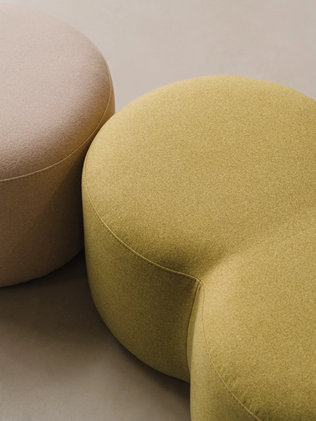 noo.ma Umbo Pouf, a set of two organic modern poufs, one in a soft beige and the other in a muted olive green, upholstered in a subtly textured, durable fabric and featuring a distinctive kidney-bean or amoeba-like sculptural shape. These contemporary, low-profile accent ottomans offer versatile seating or footrest options, perfect for minimalist living rooms, Scandinavian bedrooms, or organic modern lounge areas.