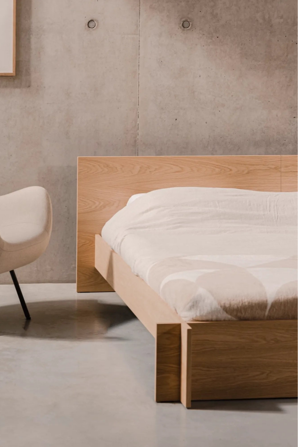 noo.ma Valt Bed, a minimalist modern bed frame crafted from light natural wood with a sleek, low-profile design and an integrated rectangular headboard. The clean lines and warm wood grain perfectly complement the soft white and beige patterned bedding, creating a serene sleeping space. Ideal for Scandinavian bedrooms, japandi interiors, or contemporary minimalist homes.
