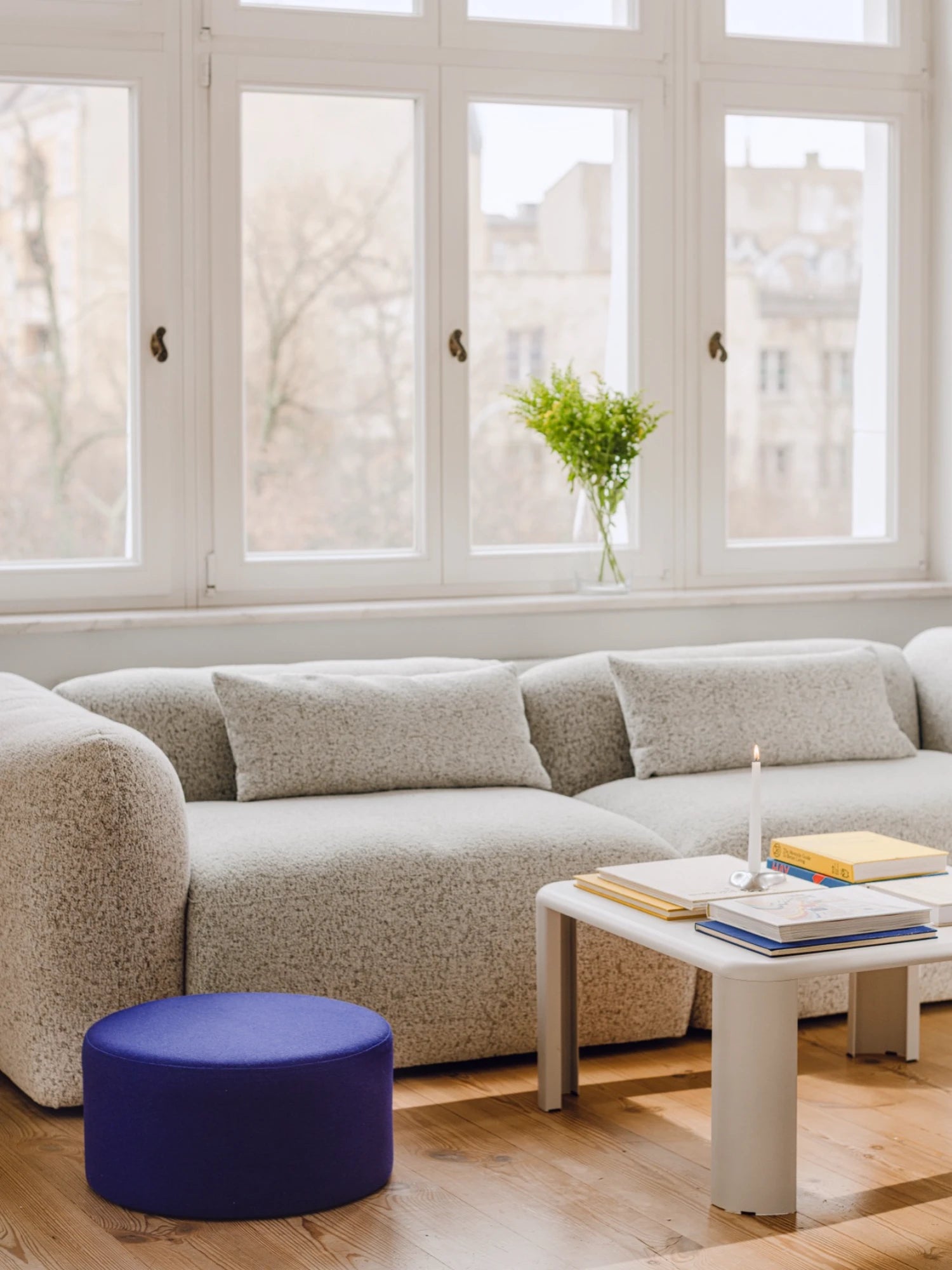Modern living room with a beige sectional sofa, blue ottoman, and a small table with books and a candle.