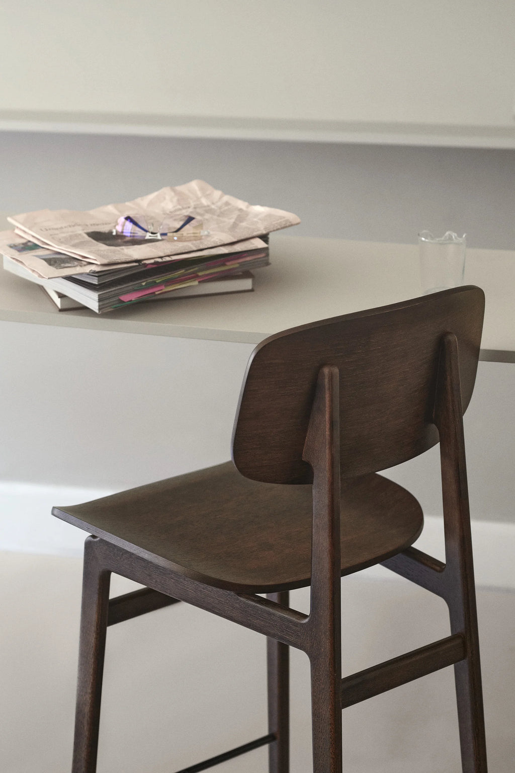 NORR11 NY11 Counter Chair in dark stained oak, seen from behind, with a matching wooden seat, positioned at a light grey counter with stacked magazines and a glass, creating a cozy, modern study nook.