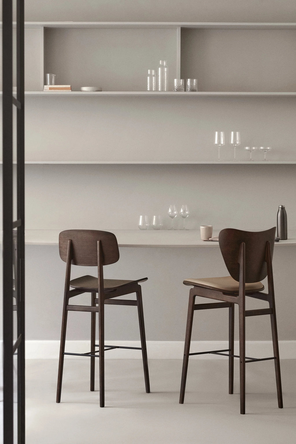 Two NORR11 NY11 Counter Chairs in dark stained oak, one with a light upholstered seat, positioned at a light grey counter with various glassware and a thermos, showcasing a sophisticated kitchen or bar area.