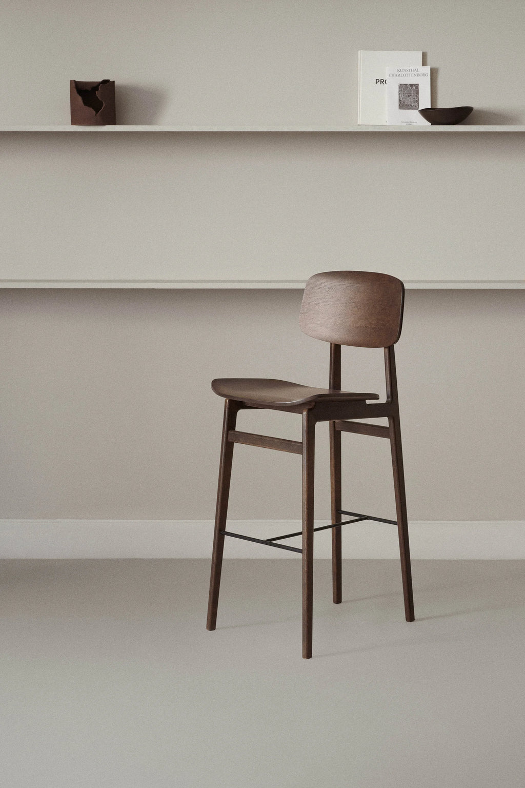 NORR11 NY11 Counter Chair in dark stained oak with a matching wooden seat, elegantly placed in a minimalist room with light grey walls and shelves adorned with decorative items, highlighting its versatile design.