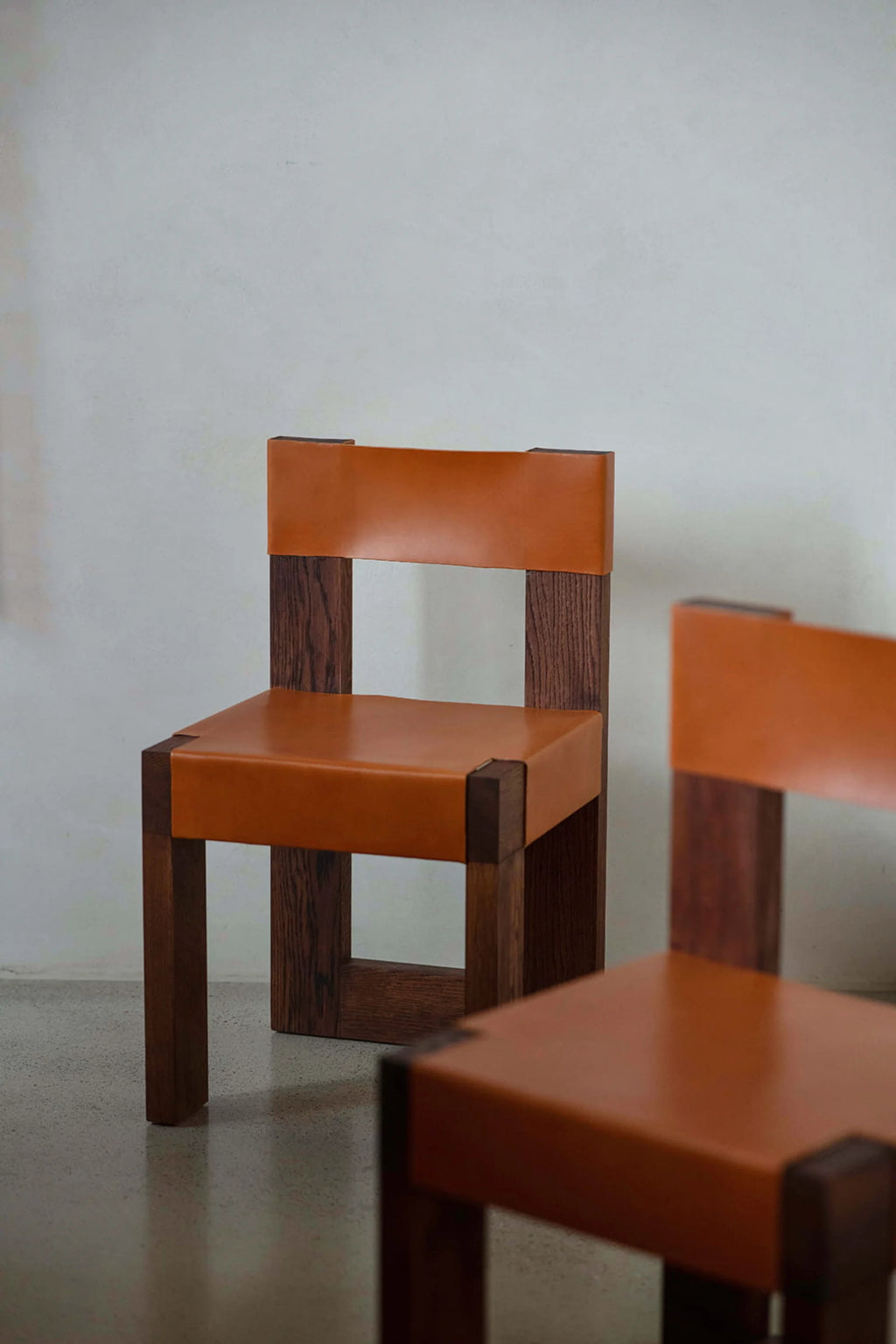 NG DESIGN Object 118 Chair, a luxury mid-century modern style wood chair with a distressed dark wood frame and rich tan leather seat and back, photographed against an off-white wall. Ideal for contemporary dining or accent seating.