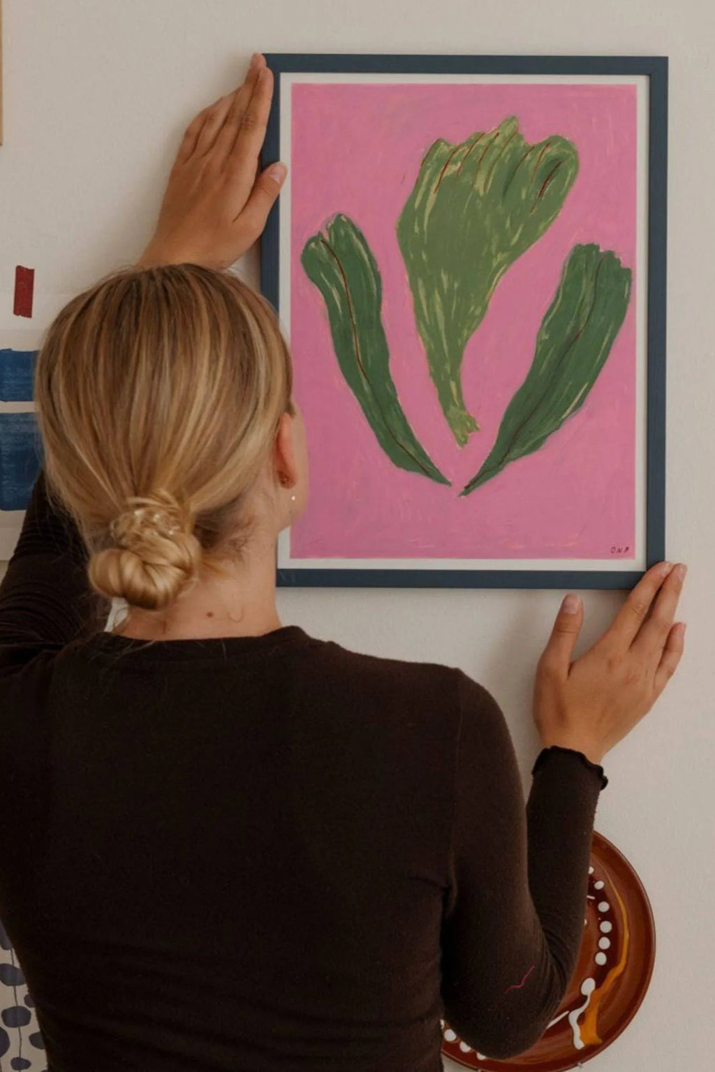 The Poster Club Danser Omkring Brombærkrat, a vibrant abstract art print featuring bold olive green organic leaf-like shapes on a bright pink background, presented in a sleek dark blue frame. This contemporary poster is perfect as modern wall art for Scandinavian living rooms, a colorful addition to a gallery wall, or a statement piece in creative studio spaces.