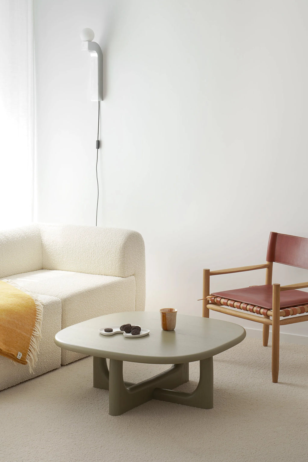 TAMO Olfo Coffee Table, a modern, organic-shaped sage green wooden table with a unique X-shaped base, styled with cookies and a mug in a neutral living room setting. A premium statement piece for any sophisticated living space.